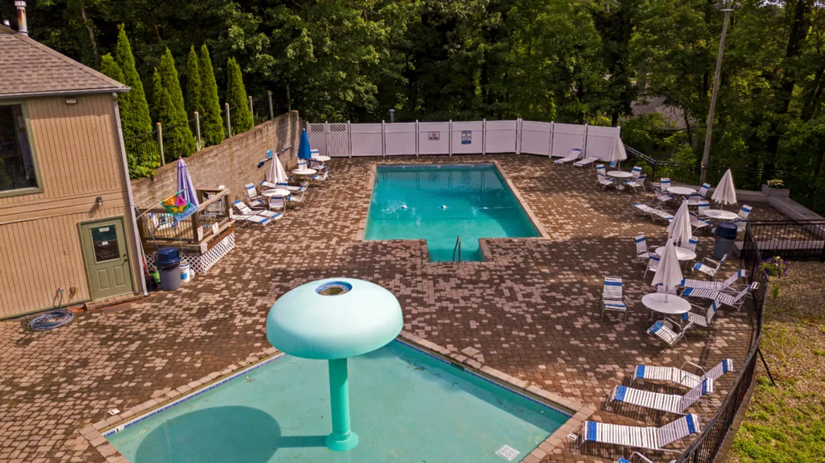 A vibrant exterior view of a swimming pool with a water feature, surrounded by lounge chairs and lush greenery. Groups of friends gather at the poolside, while children play in the water, conveying both relaxation and social enjoyment.