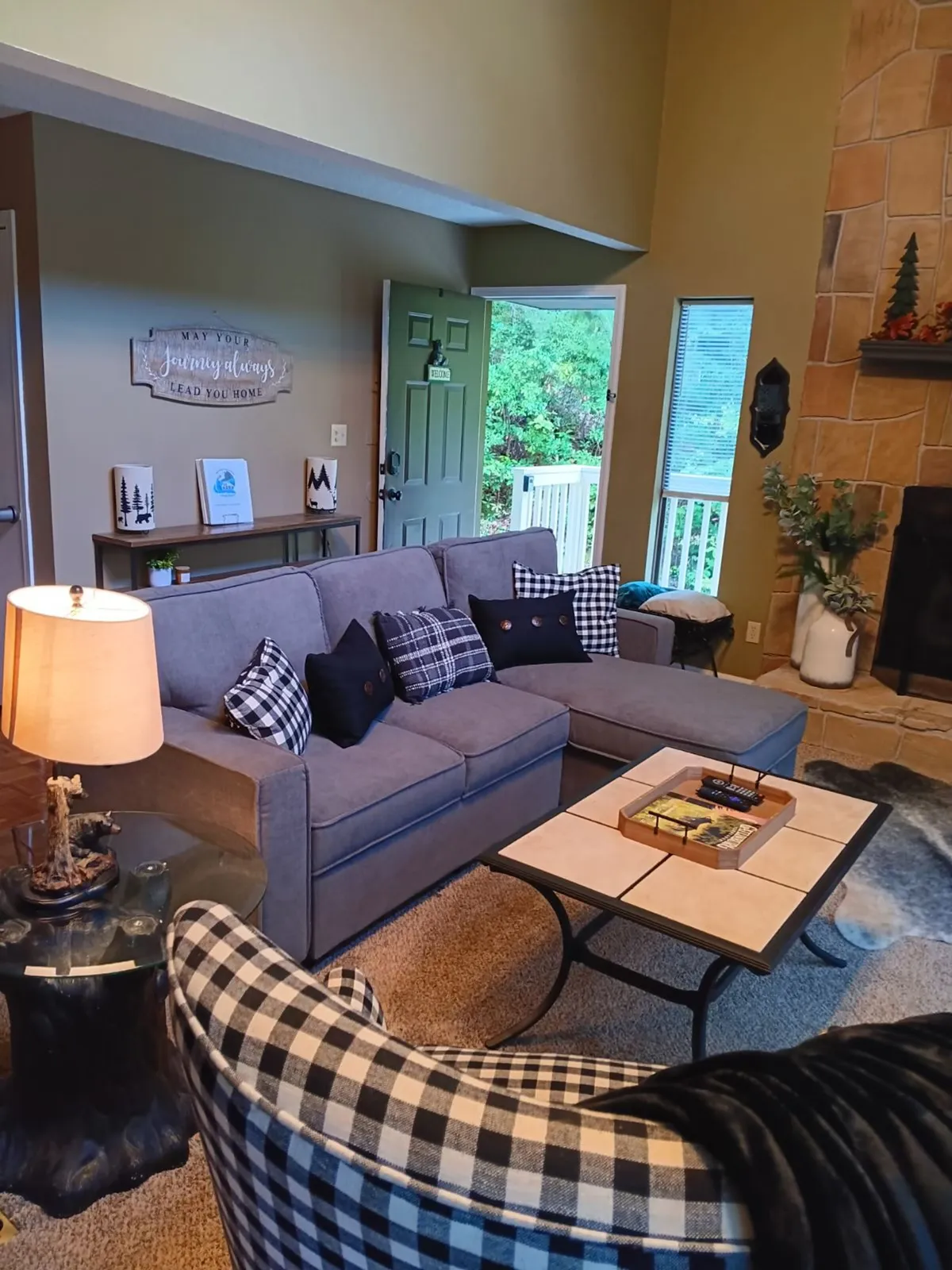 A cozy living room with a sofa and accent chairs arranged for conversation, complemented by a coffee table and decorative artwork on the walls. Large windows in the background bring in natural light and offer views of the forest beyond, enhancing the tranquil setting.