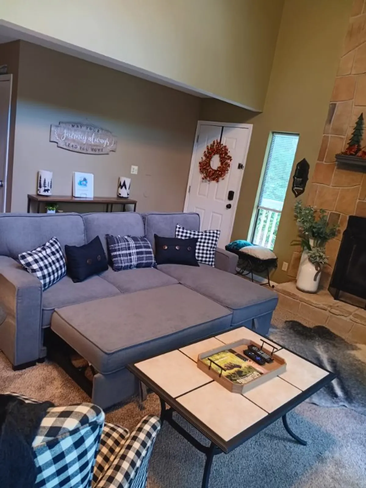 A warmly lit living room with a sectional sofa arranged around a stone fireplace. Decorative touches like a wreath above the mantel and a wooden coffee table add to the inviting ambiance. Large windows in the background reveal glimpses of the tranquil forest outside, and plush seating creates a cozy spot for relaxation or intimate conversation.