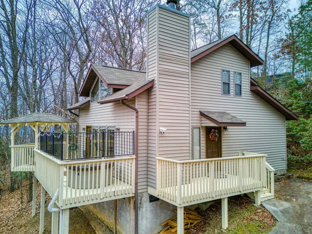 The image showcases a charming mountain chalet nestled among tall trees, with a spacious wooden deck and a distinctive gabled roof. The chimney hints at cozy evenings, while the serene forest setting bathes the scene in warm, peaceful light—suggesting sunset or sunrise. The deck is inviting for morning coffee or unwinding as the sun sets over the Smoky Mountains.