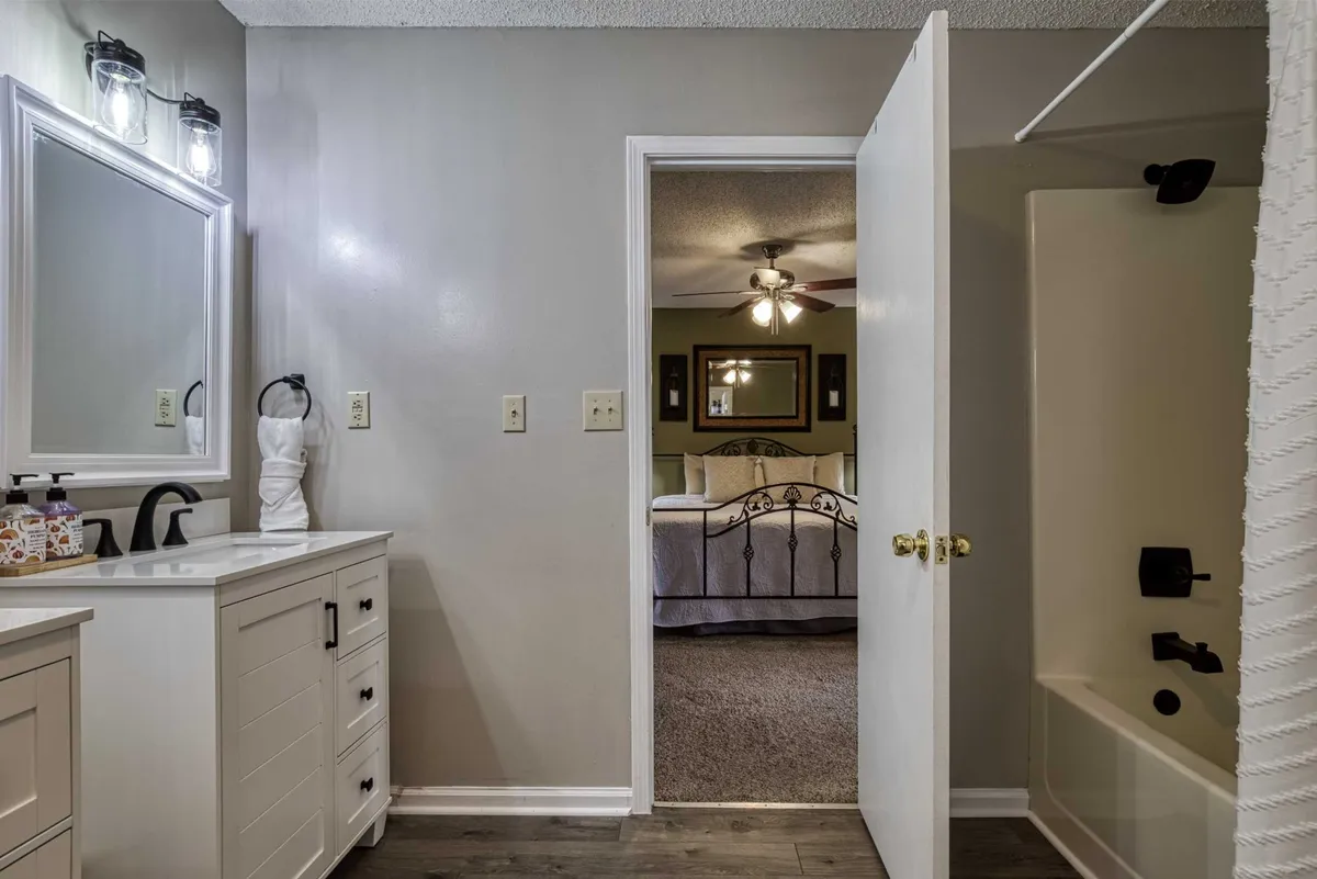 A functional and serene bathroom space with a vanity and mirror, partially open door leading into the bedroom, and a bathtub inviting a relaxing soak. The scene exudes a cozy, private atmosphere ideal for unwinding.